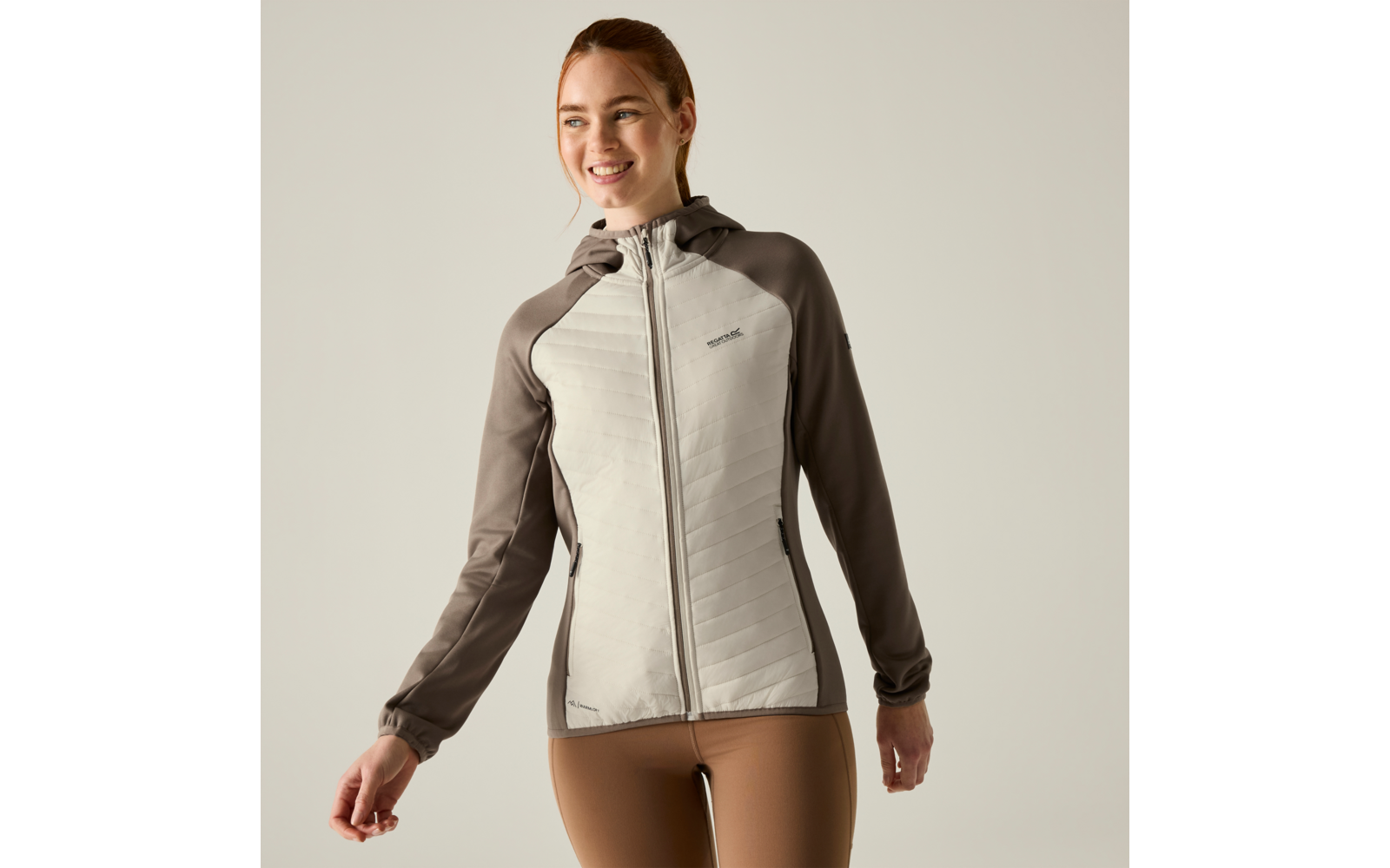 Person standing against a light background, wearing a light grey quilted hooded jacket with darker sleeves and beige leggings, looking to the side and smiling