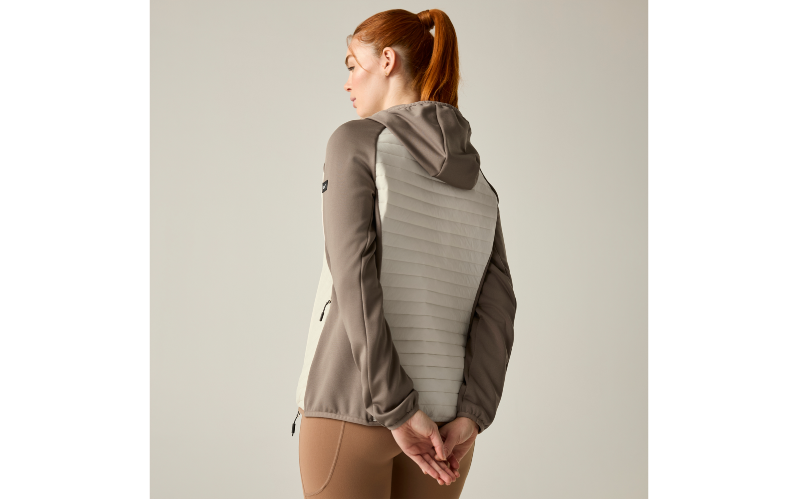 Rear view of a woman with a red ponytail wearing a hooded jacket with a light quilted back, hands clasped behind her back