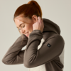 Person with red hair in a ponytail, side view, smiling, pulling up the hood of a brown hoodie; plain light background