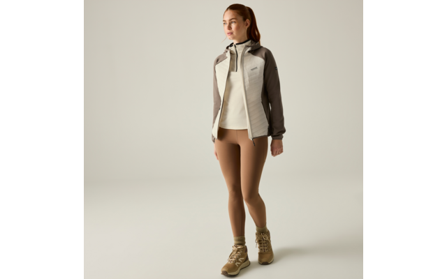 Person standing on a light background, wearing a light beige-grey hooded jacket, light sweater, brown leggings and beige hiking boots, hair in a ponytail