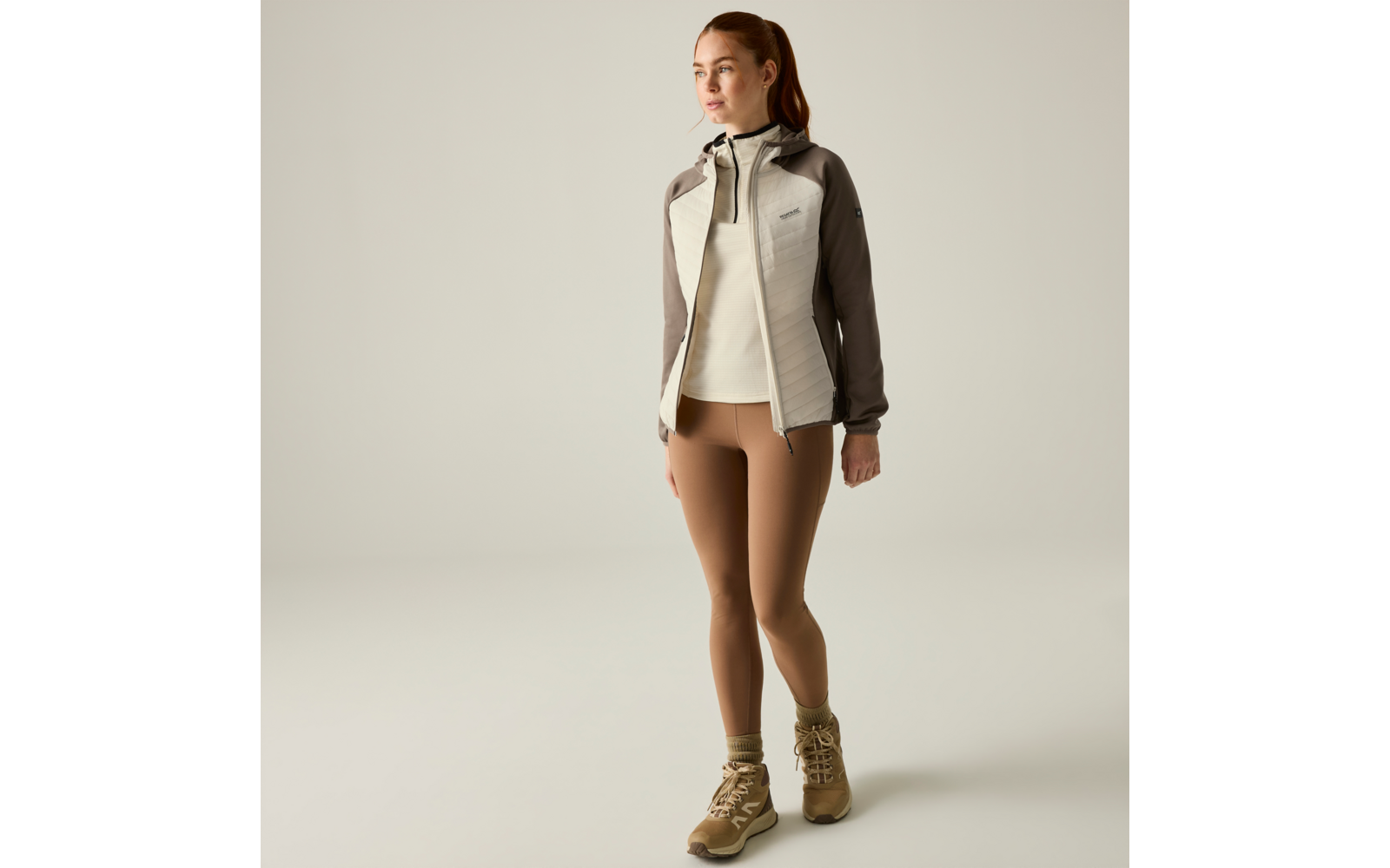 Person standing on a light background, wearing a light beige-grey hooded jacket, light sweater, brown leggings and beige hiking boots, hair in a ponytail