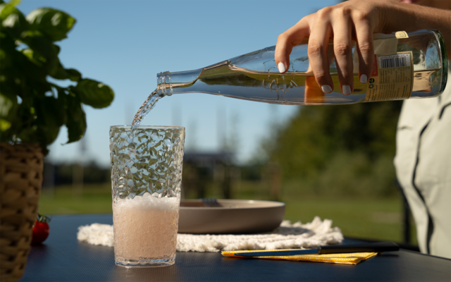 Hand giet vloeistof uit een fles in een glas met schuim op een buitentafel; daarnaast een potplant, bord, doek en pen; vage bomen en blauwe lucht op de achtergrond