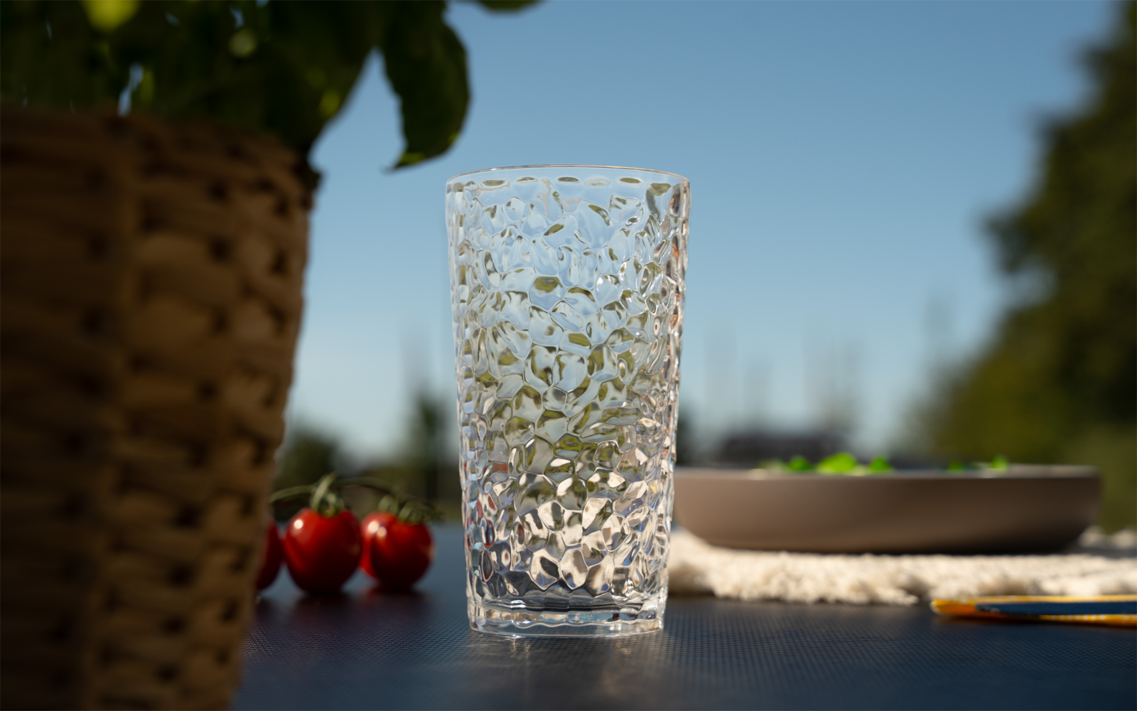 Durchsichtiges, strukturiertes Trinkglas auf dunklem Tisch; links geflochtener Blumentopf und zwei Kirschtomaten; rechts unscharfe Schale mit grünen Objekten; unscharfer blauer Himmel im Hintergrund