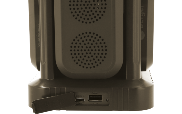 Parte inferior de un dispositivo negro con dos rejillas de altavoz redondas, soportes verticales, puertos visibles (USB‑C y ranura) y una tapa de goma colgando