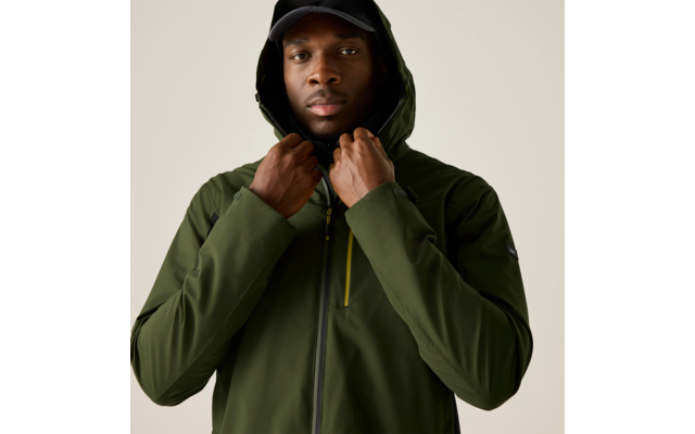 Person trägt grüne Kapuzenjacke und schwarze Kappe, hält mit beiden Händen den Kragen, Blick zur Kamera, neutraler Hintergrund