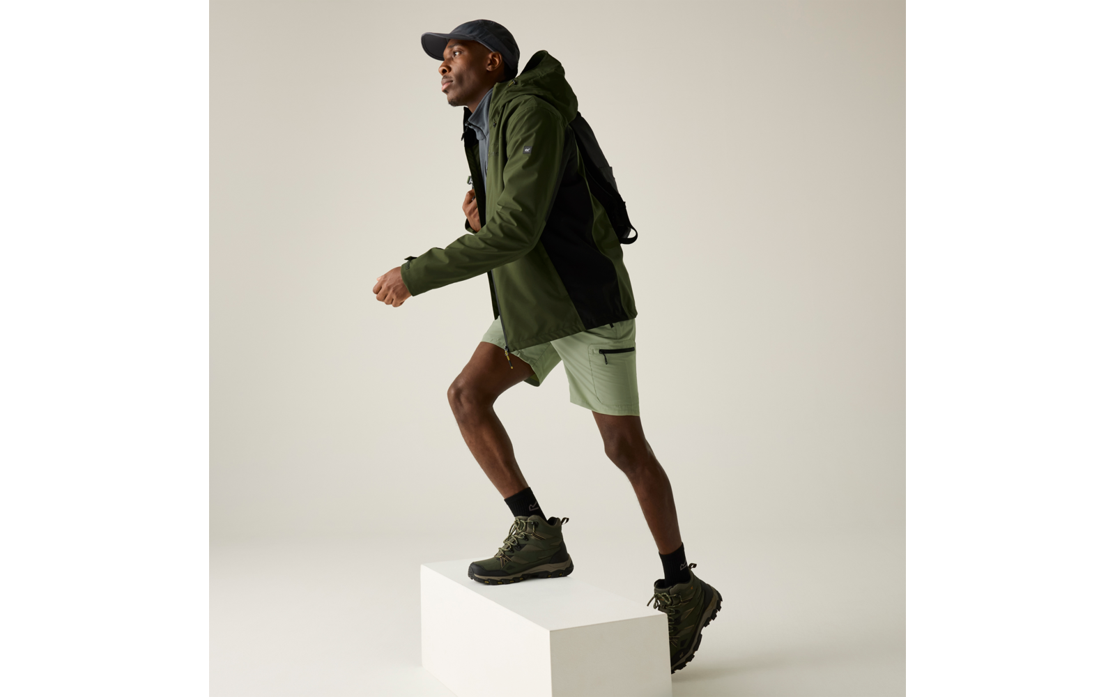 Person trägt grüne Jacke, hellgrüne Shorts, schwarze Kappe und schwarzen Rucksack, schwarze Socken und grüne Wanderschuhe, mit einem Fuß auf einem weißen Podest vor neutralem Hintergrund