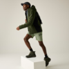 Person trägt grüne Jacke, hellgrüne Shorts, schwarze Kappe und schwarzen Rucksack, schwarze Socken und grüne Wanderschuhe, mit einem Fuß auf einem weißen Podest vor neutralem Hintergrund