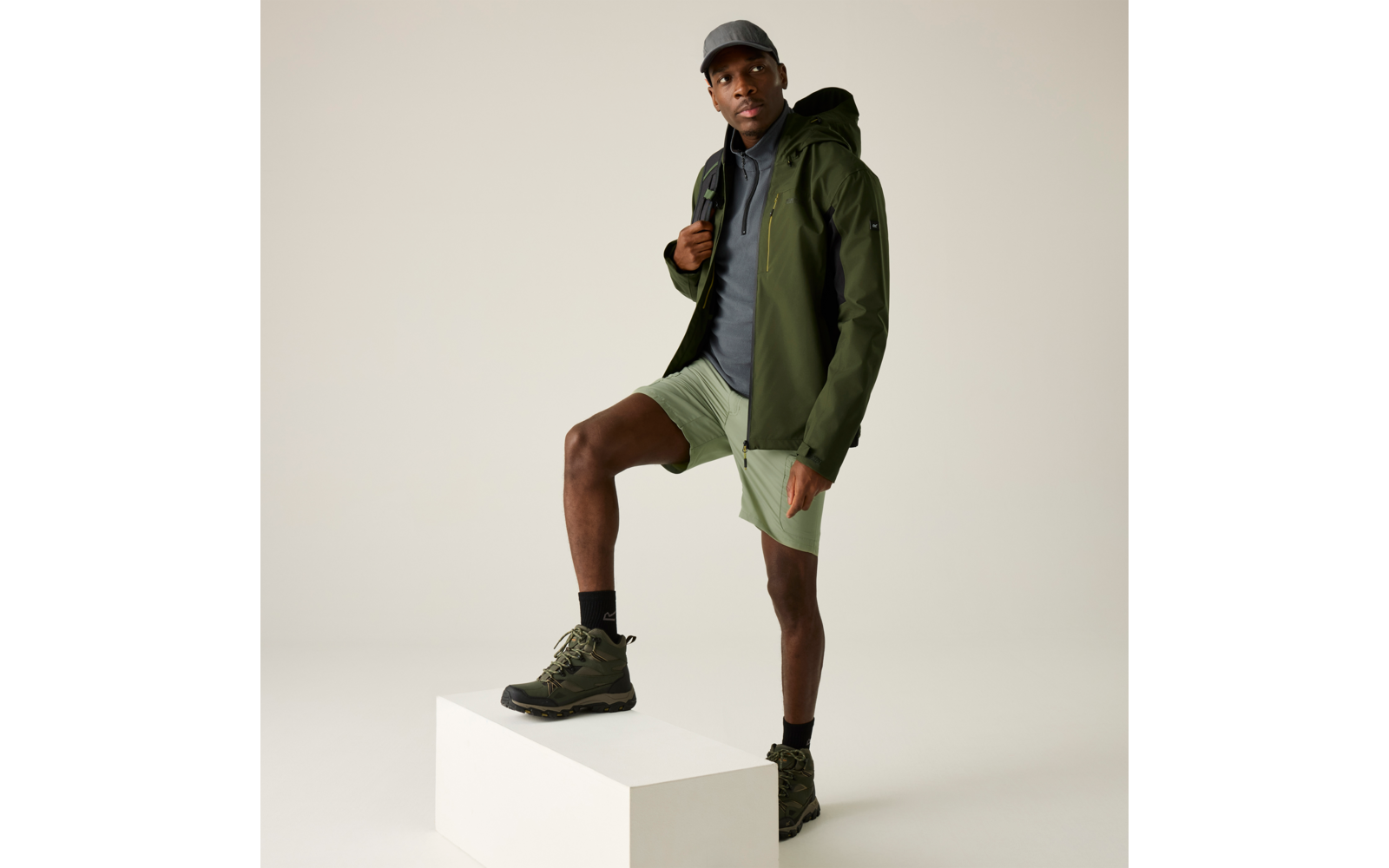 Person steht mit einem Fuß auf einem weißen Podest, trägt olivgrüne Kapuzenjacke, graues Kapuzenshirt, hellgrüne Shorts, graue Kappe, schwarze Socken und olivgrüne robuste Schuhe; eine Hand hält den Jackenkragen; einfarbiger heller Hintergrund