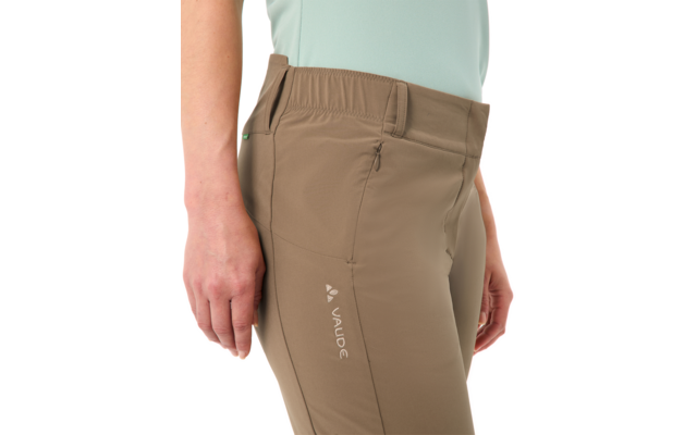 Nahaufnahme der Hüft- und Oberschenkelpartie einer Person mit hellbraunen Wanderhosen mit Reißverschlusstasche, sichtbarem VAUDE-Logo, elastischem Bund und hellblauem Oberteil; Hände seitlich