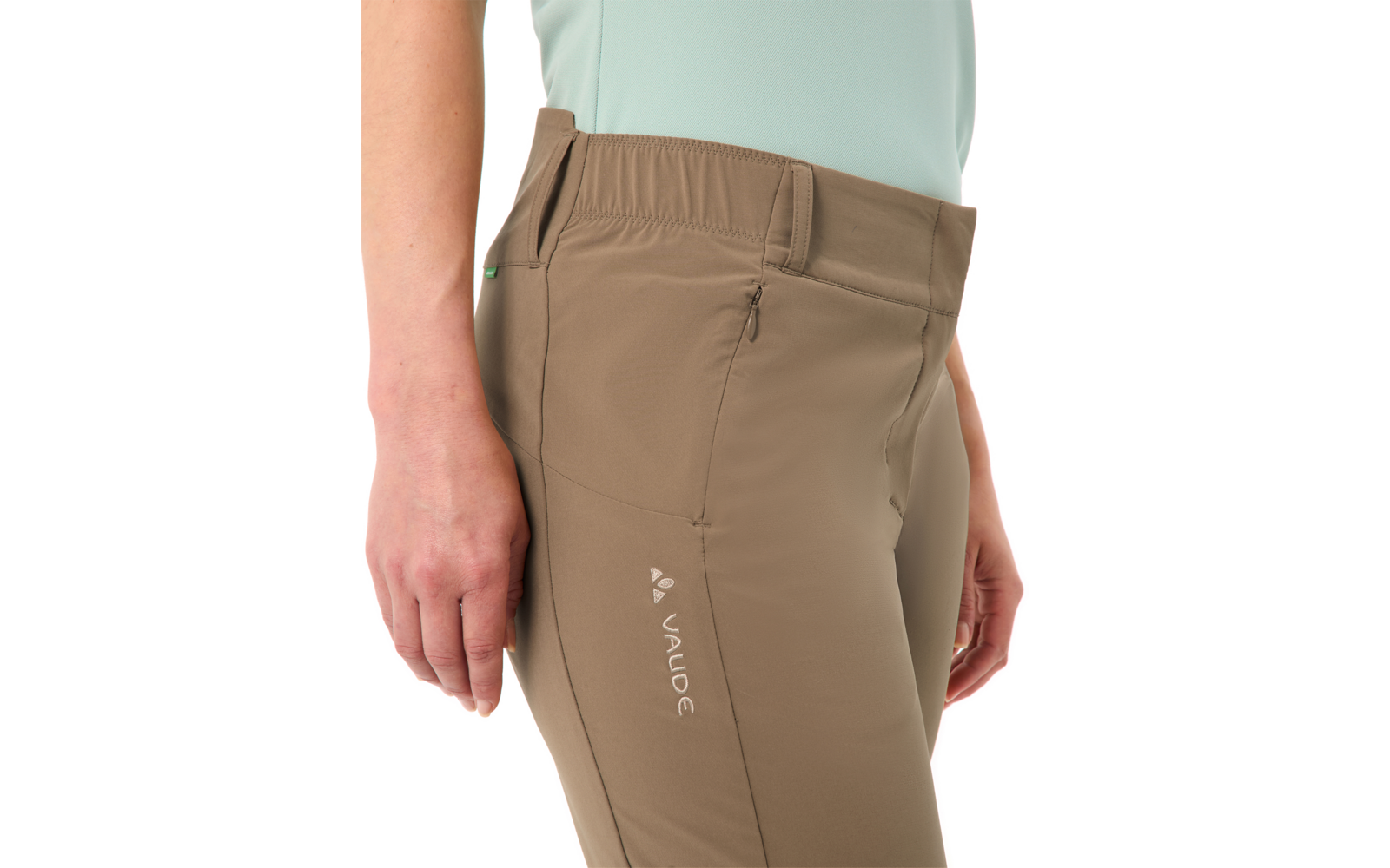 Nahaufnahme der Hüft- und Oberschenkelpartie einer Person mit hellbraunen Wanderhosen mit Reißverschlusstasche, sichtbarem VAUDE-Logo, elastischem Bund und hellblauem Oberteil; Hände seitlich