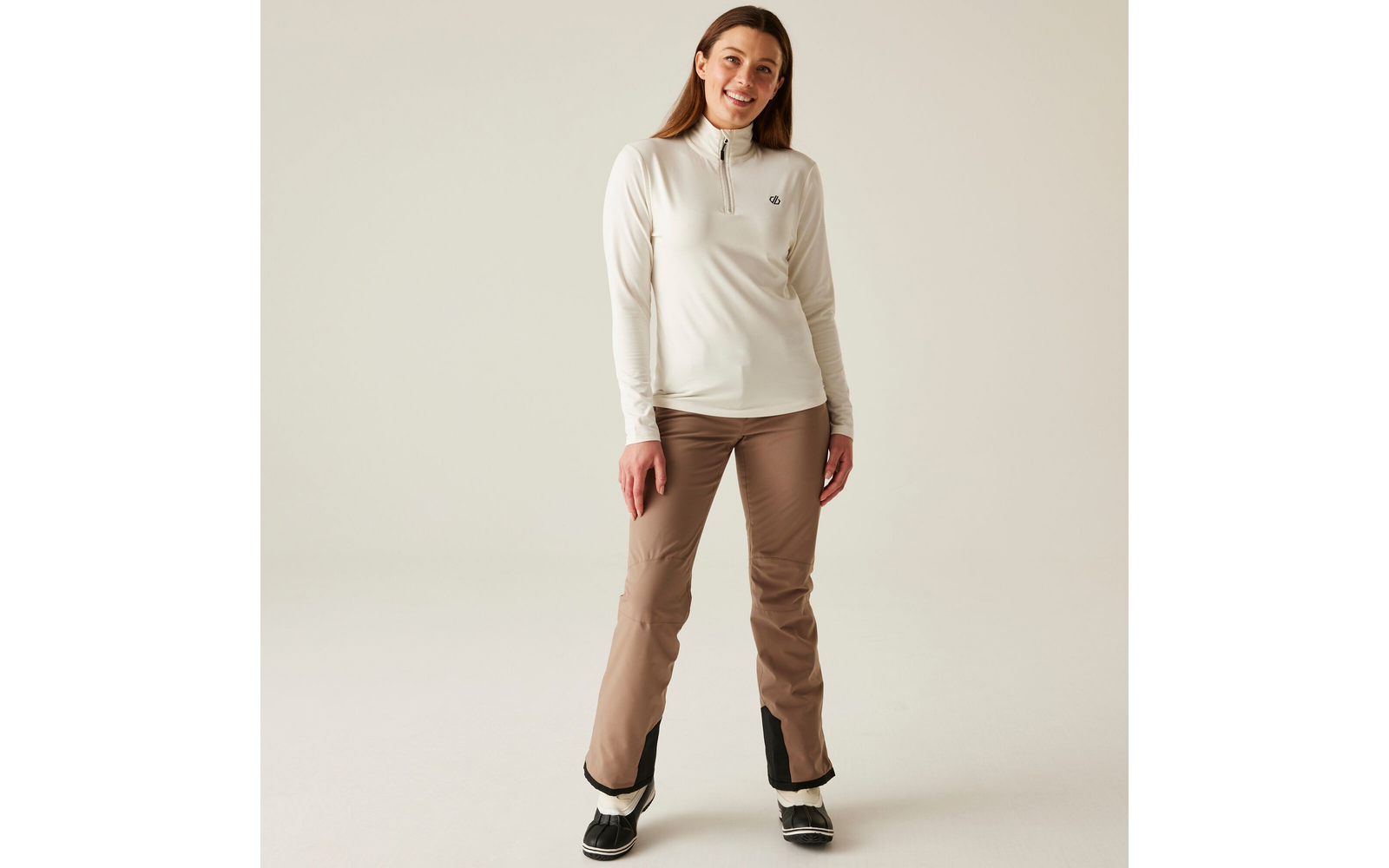 Femme debout devant un fond clair, porte une veste sweat claire et un pantalon beige
