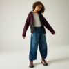 Person mit lockigen Haaren und Brille, steht auf hellem Hintergrund, trägt weißes T‑Shirt, weite blaue Jeans, dunkelviolette Jacke mit schwarzen Einsätzen und rosa Turnschuhe