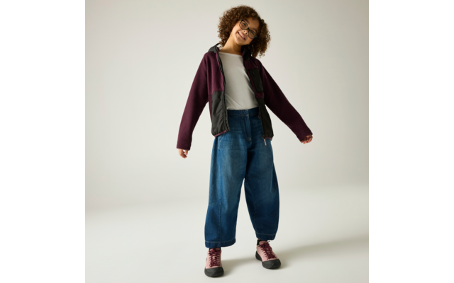 Person mit lockigen Haaren und Brille, steht auf hellem Hintergrund, trägt weißes T‑Shirt, weite blaue Jeans, dunkelviolette Jacke mit schwarzen Einsätzen und rosa Turnschuhe