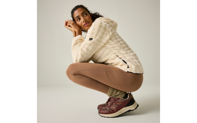 Persona agachada, viste chaqueta polar crema con capucha, mallas marrones, calcetines verdes y zapatillas burdeos, fondo claro