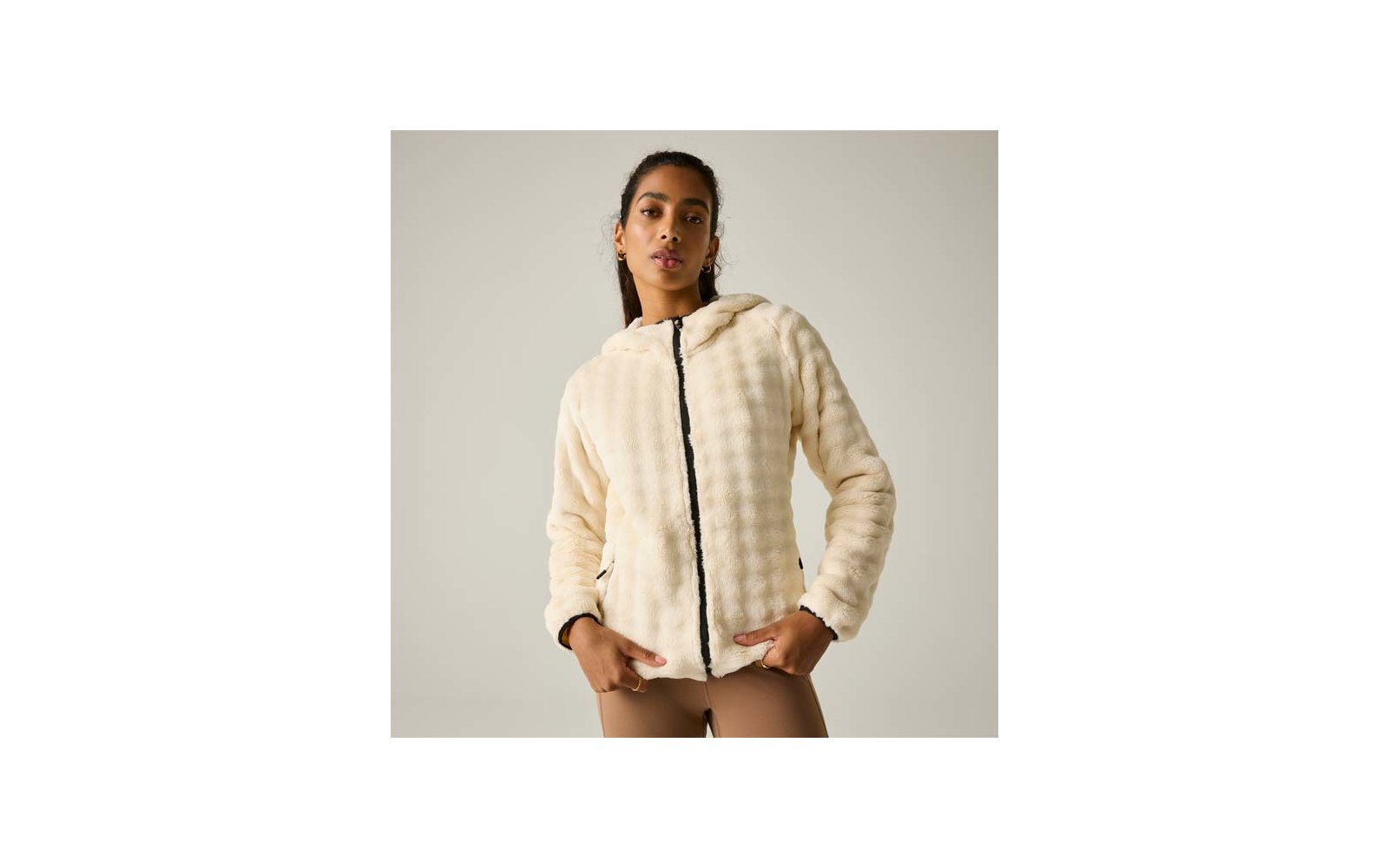 Persona con el cabello recogido que lleva una chaqueta con capucha de pelo sintético color crema con cremallera negra y leggings marrón claro sobre fondo claro