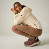 Persona agachada, viste chaqueta polar crema con capucha, mallas marrones, calcetines verdes y zapatillas burdeos, fondo claro