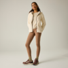 Persona de pie sobre fondo claro, lleva chaqueta polar crema, camiseta clara, mallas marrones, calcetines verdes y zapatillas burdeos, manos en los bolsillos