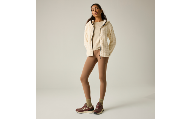 Persona de pie sobre fondo claro, lleva chaqueta polar crema, camiseta clara, mallas marrones, calcetines verdes y zapatillas burdeos, manos en los bolsillos