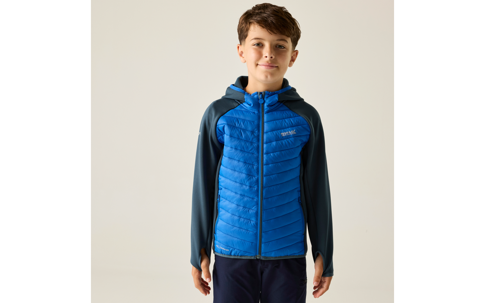 Junge steht vor hellem Hintergrund, trägt blau gesteppte Jacke mit Kapuze, Hände seitlich, blickt in die Kamera