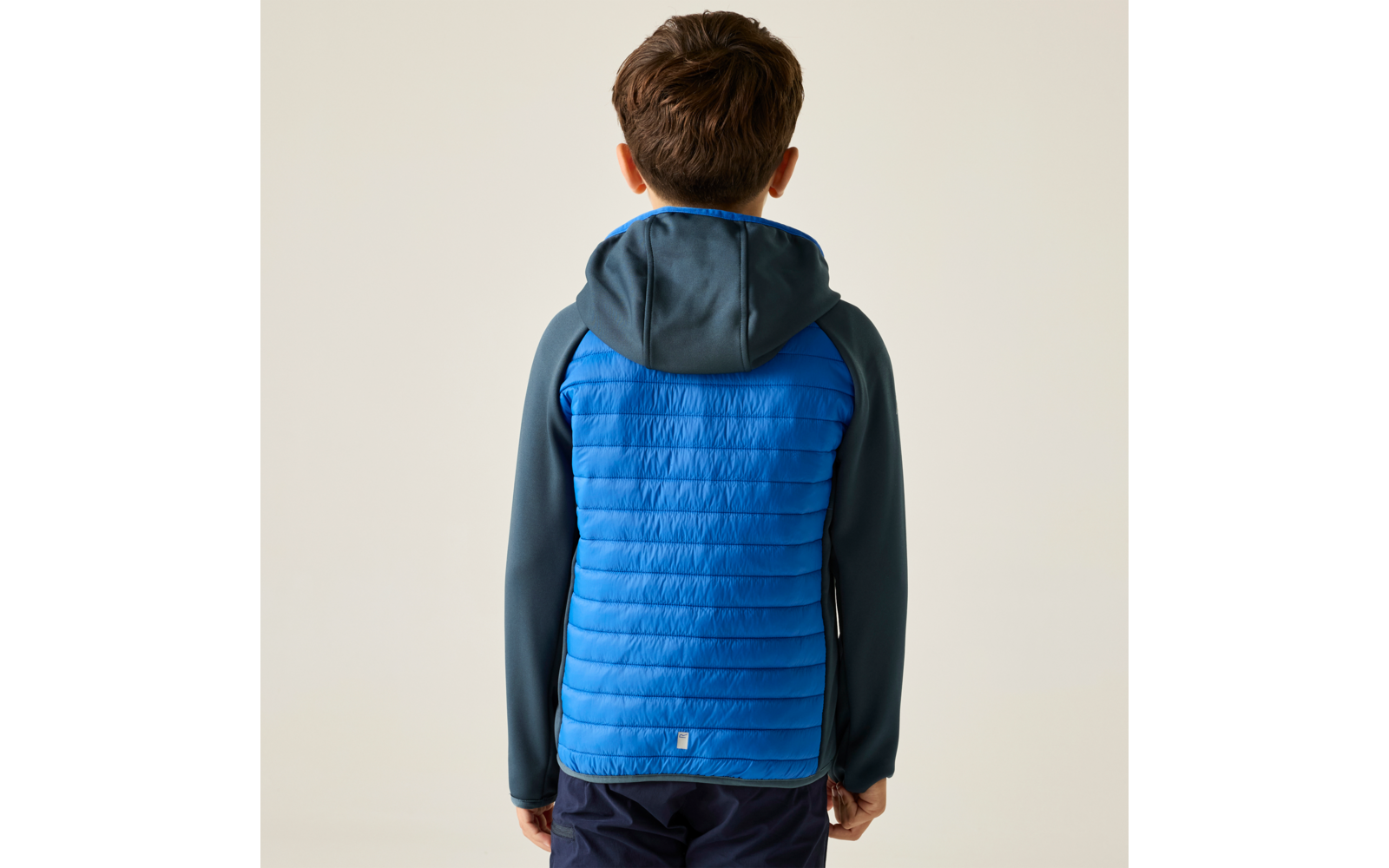 Rückenansicht einer Person mit kurzem braunen Haar, trägt eine blaue gesteppte Jacke mit dunkelblauen Ärmeln und Kapuze, dunkle Hosen, heller einfarbiger Hintergrund