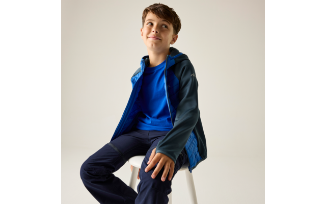 Junge sitzt auf weißem Hocker, trägt blaue Jacke und dunkle Hose, blickt nach oben, heller einfarbiger Hintergrund