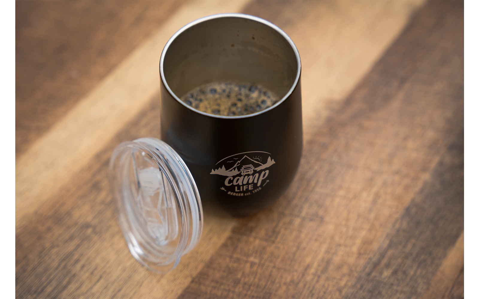 Een zwarte beker met koffie op een houten tafel, naast een doorzichtige deksel