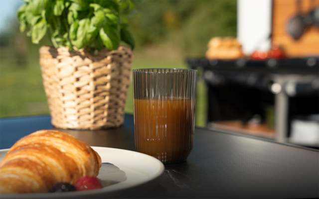 Rillenglas mit braunem Getränk auf einem Tisch, vorne ein Teller mit Croissant und zwei Beeren, links Basilikum in geflochtenem Topf, im Hintergrund unscharfer Grill