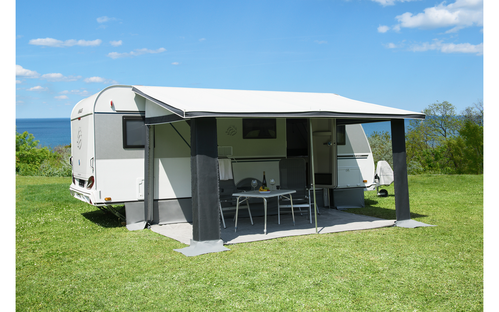 Weißer Wohnwagen auf grüner Wiese, ausgeklappte Markise, darunter ein Tisch mit zwei Stühlen, im Hintergrund Meer und blauer Himmel mit Wolken