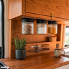 Houten kast en werkblad; drie hangende glazen potten met zwarte deksels (donkere deeltjes, witte deeltjes, oranje poeder); zwarte pot met groene plant; twee messen op magneetstrip; klein gevlochten mandje; keramische mok; raam met onscherp buitenzicht; metalen sluiting aan kast; stopcontact