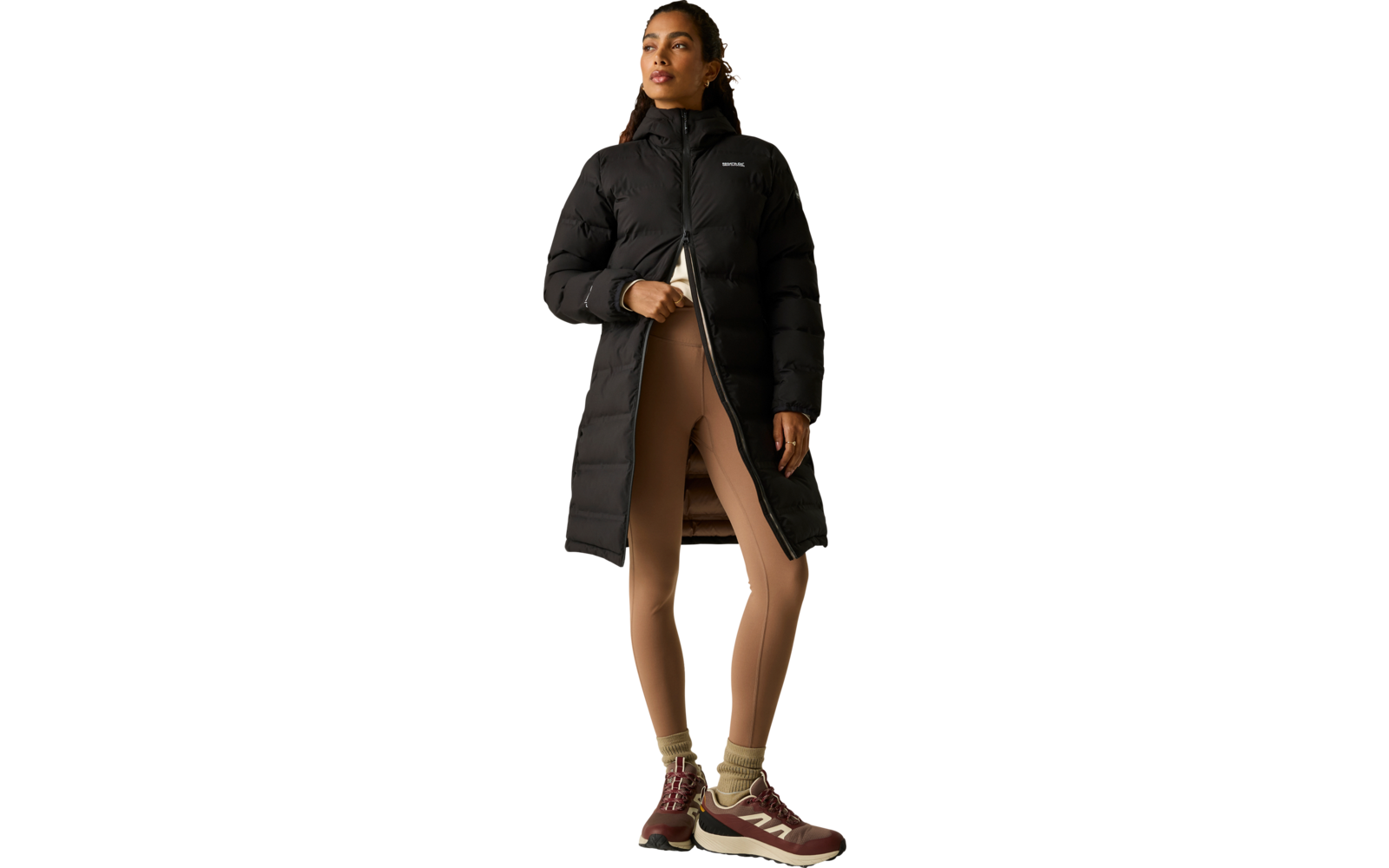 Persona con cappotto imbottito nero lungo, leggings beige e scarpe da ginnastica marroni, in piedi con una mano sulla cerniera, sguardo in alto a destra, sfondo chiaro
