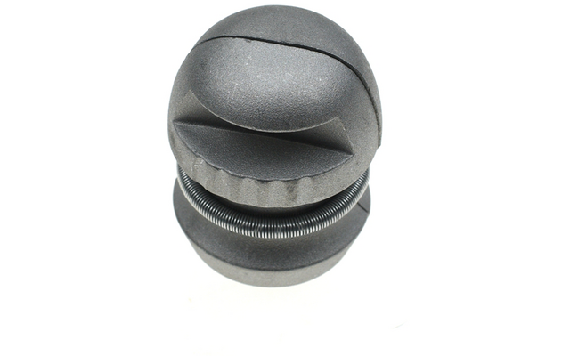 Graues zylindrisches Objekt aus zwei gestapelten, abgerundeten Scheiben; obere Scheibe mit gebogener Schlitzöffnung; zwischen den Scheiben sichtbare Metallspiralfeder; weißer Hintergrund