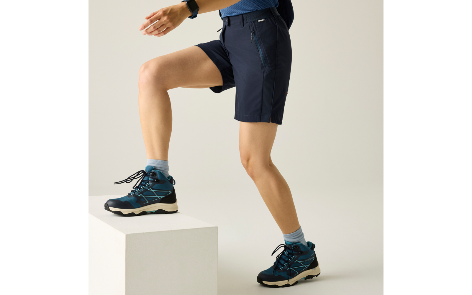 Unterkörper einer Person von Hüfte bis Fuß, dunkelblaue Shorts, blaugrüne Sportschuhe, ein Fuß auf einem weißen Block; hellblaue Socken und Armbanduhr sichtbar