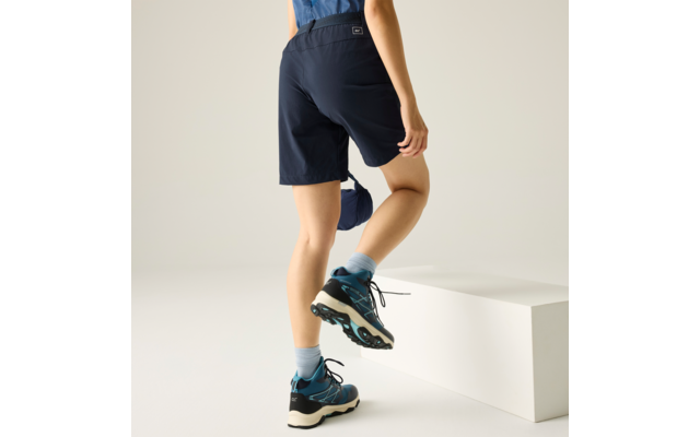 Unterschenkel und Füße einer Person in dunkelblauen Shorts, hellblauen Socken und blauen Schuhen; ein Fuß auf einem weißen Block; in der Hand ein dunkelblaues Kleidungsstück