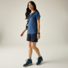 Frau in blauem T‑Shirt und dunkelblauen Shorts, blaue Sportschuhe, Umhängetasche quer, Smartwatch, vor hellem einfarbigem Hintergrund