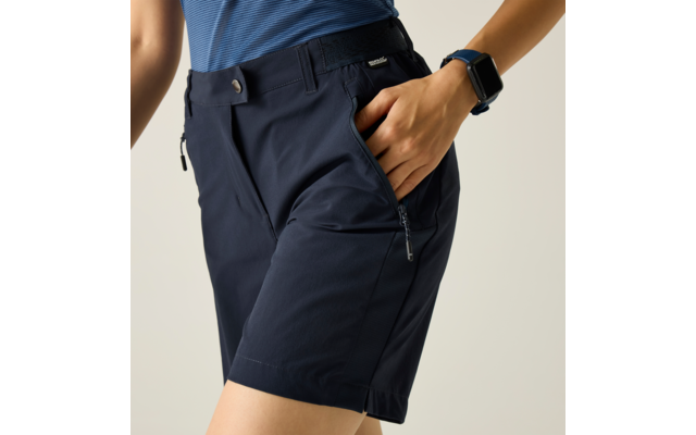 Unterer Rumpf und obere Beine einer Person, dunkelblaue Shorts mit Reißverschlusstasche, Hand in Tasche, blau gestreiftes Shirt sichtbar, Smartwatch am Handgelenk, heller Hintergrund