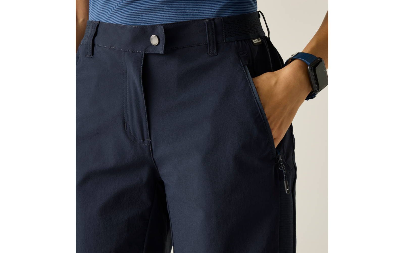 Dunkelblaue Hose mit Druckknopf und Bund, Hand in seitlicher Hosentasche, Reißverschlusstasche am Oberschenkel, blau gestreiftes Oberteil am Bund sichtbar, Smartwatch am Handgelenk