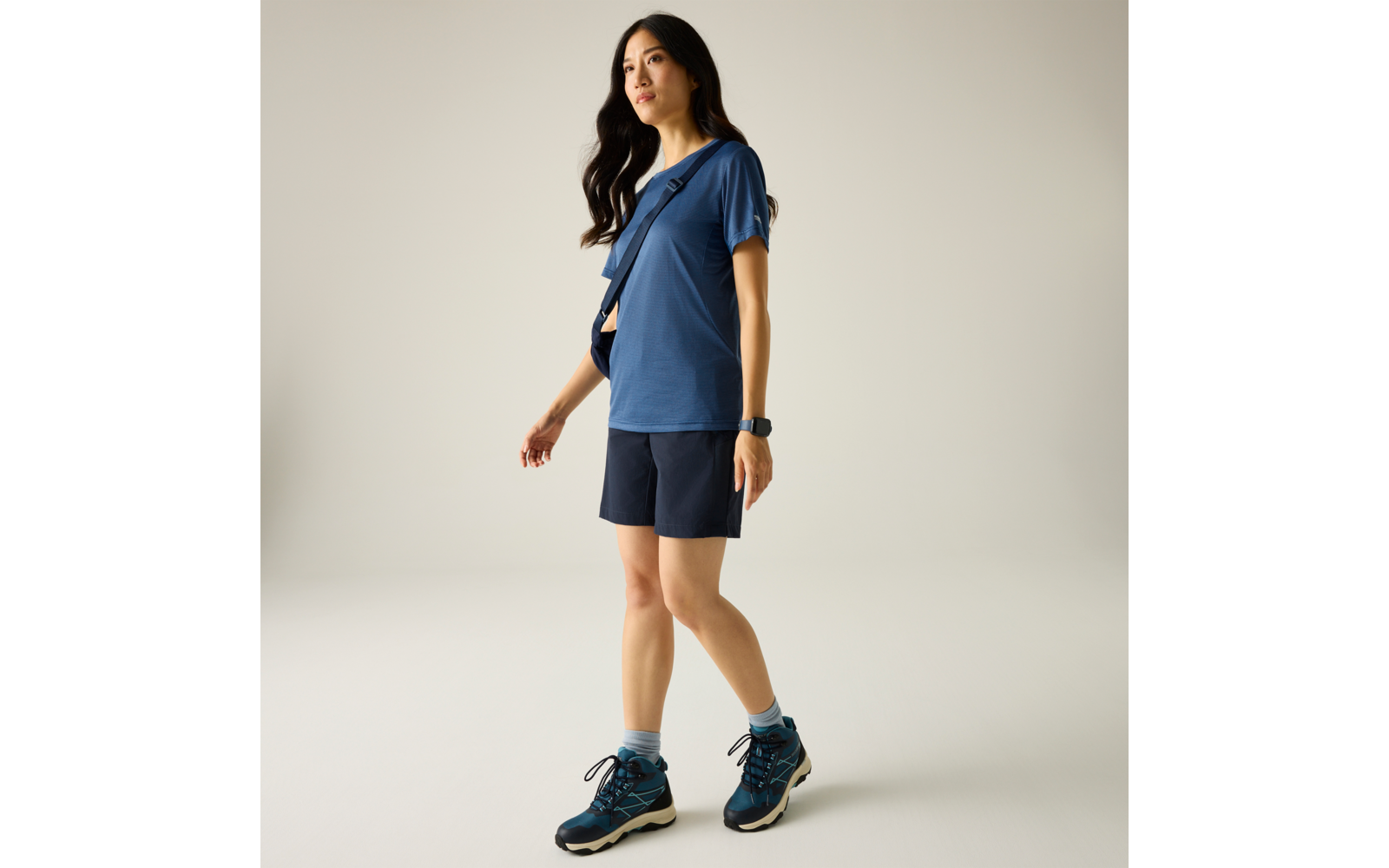Frau in blauem T‑Shirt und dunkelblauen Shorts, blaue Sportschuhe, Umhängetasche quer, Smartwatch, vor hellem einfarbigem Hintergrund