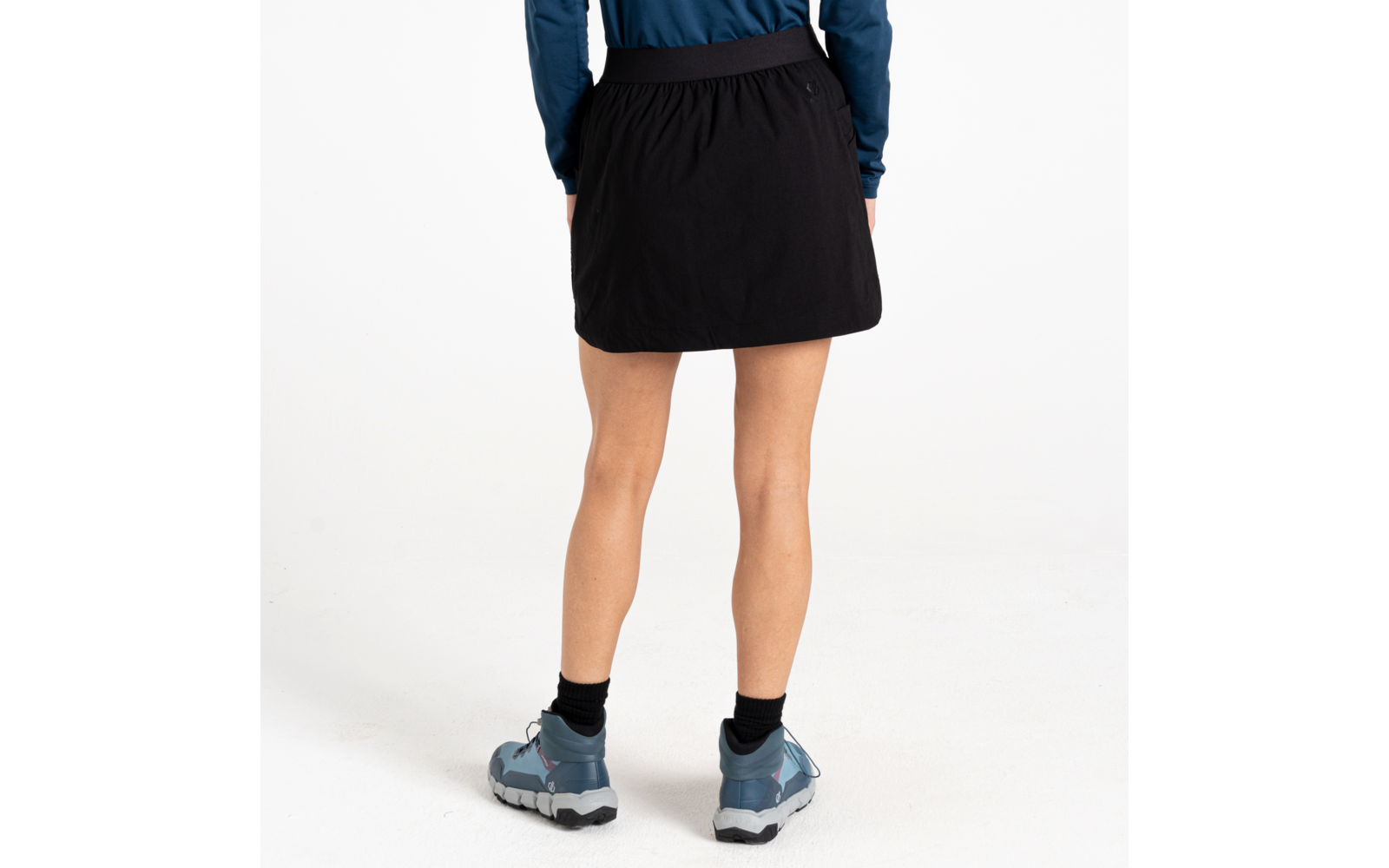 Achteraanzicht van een staande persoon met een korte zwarte rok, zwarte enkelsokken en lichtblauwe wandelschoenen op een witte achtergrond