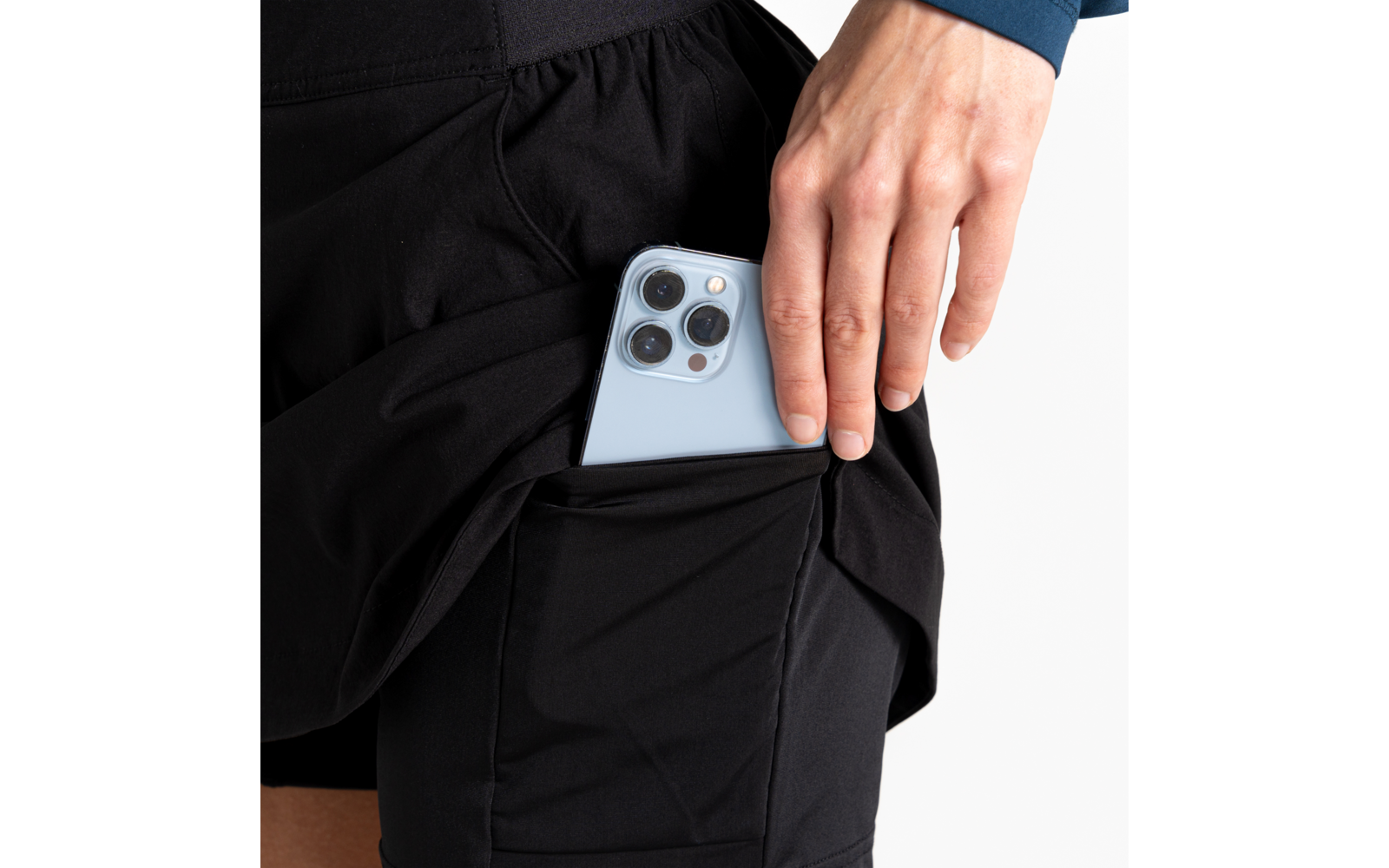 Hand houdt een lichtblauwe smartphone met drie cameralenzen in de zijzak van zwarte sportshorts, witte achtergrond