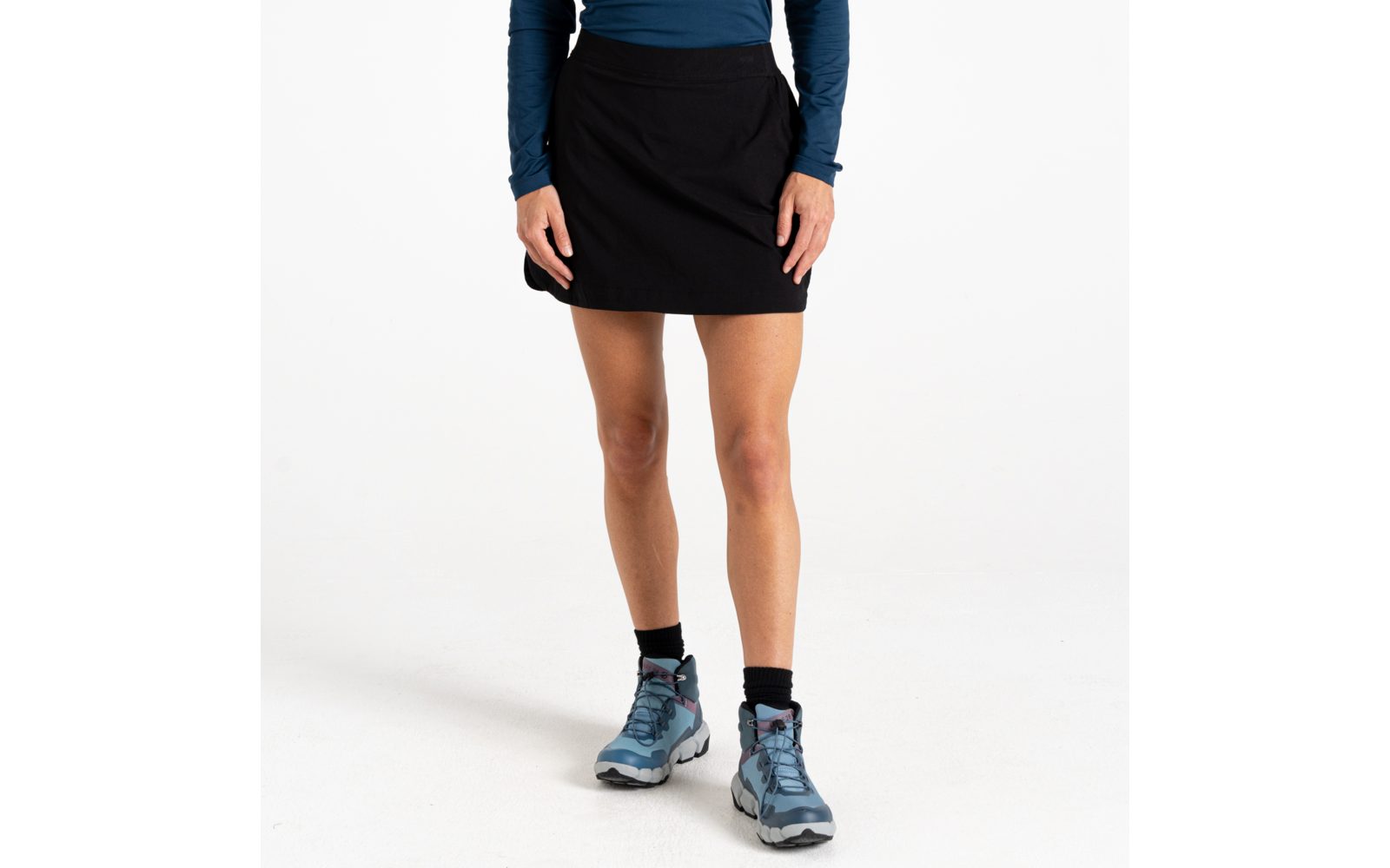 Onderlichaam van een staande persoon met een zwarte rok, blauw shirt met lange mouwen, zwarte sokken en blauwe wandelschoenen tegen een witte achtergrond