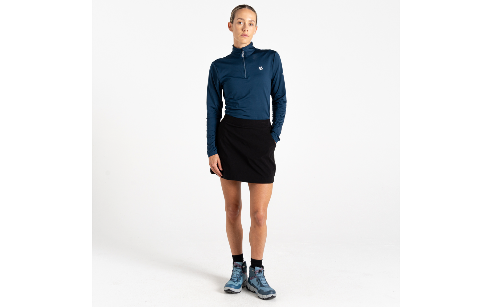 Vrouw staand, donkerblauw shirt met lange mouwen en korte rits, zwarte korte rok, enkelsokken en blauwe wandelschoenen, witte achtergrond