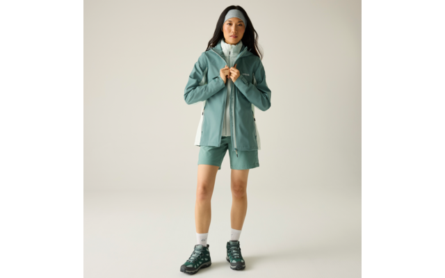 Person steht frontal, hält mit beiden Händen den Reißverschluss einer mintgrünen Outdoorjacke, trägt passende Shorts, Stirnband, weiße Knöchelsocken und dunkelgrüne Wanderschuhe vor hellem Hintergrund