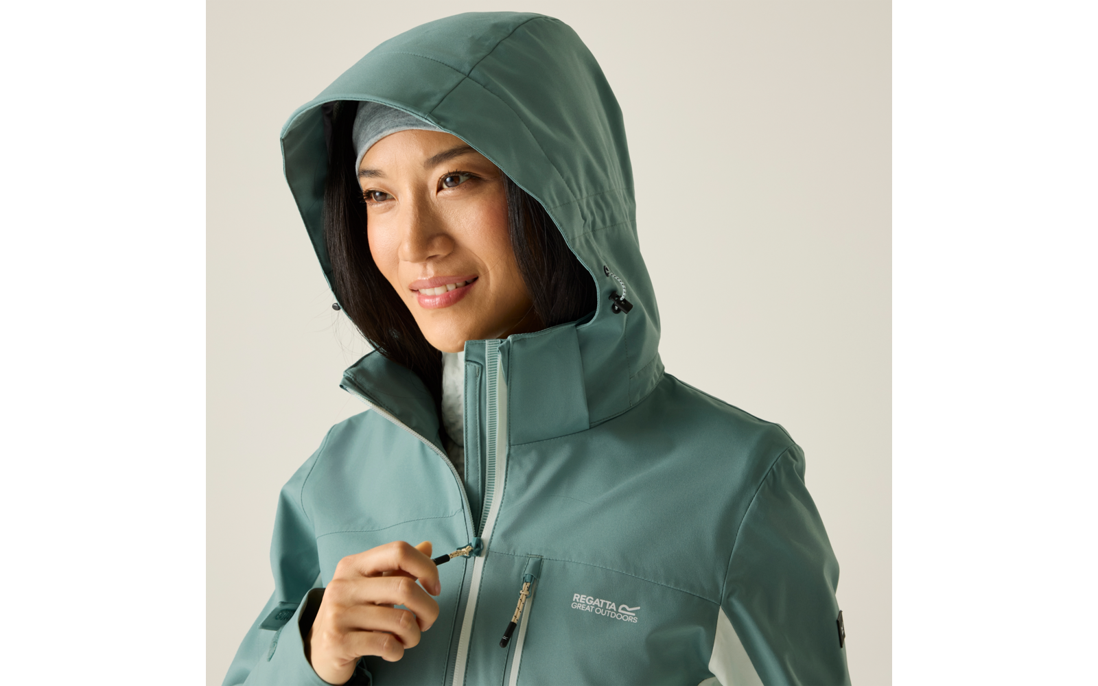 Person mit türkisgrüner Kapuzenjacke, Kapuze aufgesetzt, hält den Reißverschlusszug nahe der Brust