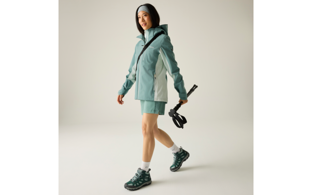 Person in mintgrüner Outdoorjacke und Shorts, mit Stirnband und Wanderschuhen, hält zusammengeklappte Wanderstöcke vor hellem Hintergrund