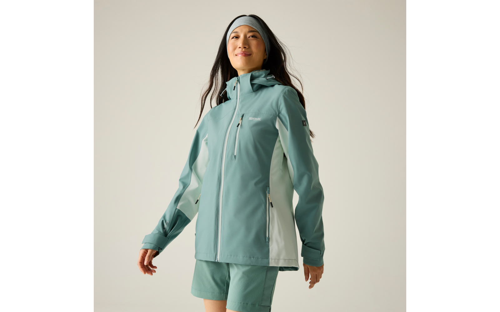 Person mit langem dunklem Haar trägt mintgrüne Outdoorjacke mit Kapuze, passende Shorts und Stirnband vor hellem Hintergrund