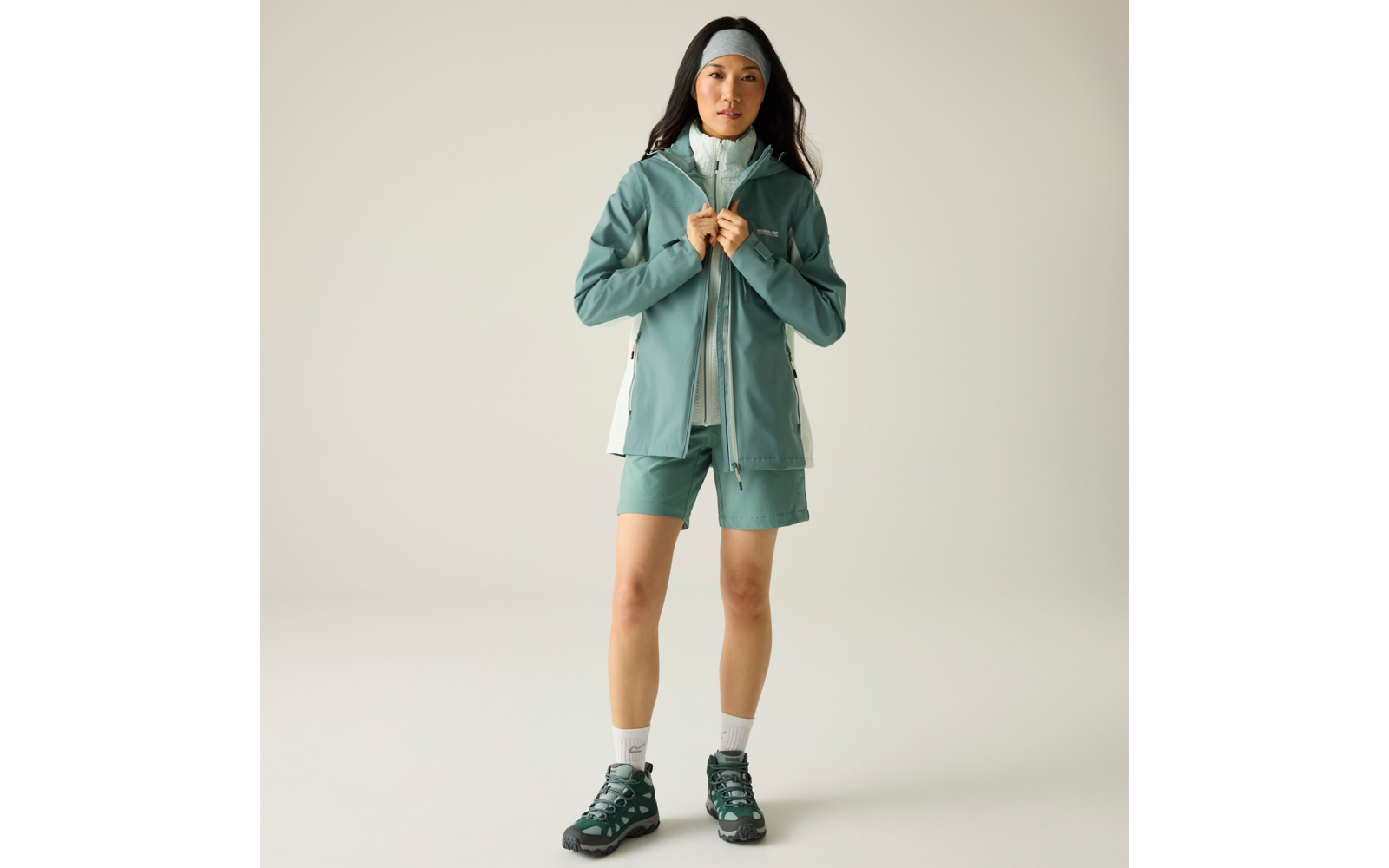 Person steht frontal, hält mit beiden Händen den Reißverschluss einer mintgrünen Outdoorjacke, trägt passende Shorts, Stirnband, weiße Knöchelsocken und dunkelgrüne Wanderschuhe vor hellem Hintergrund