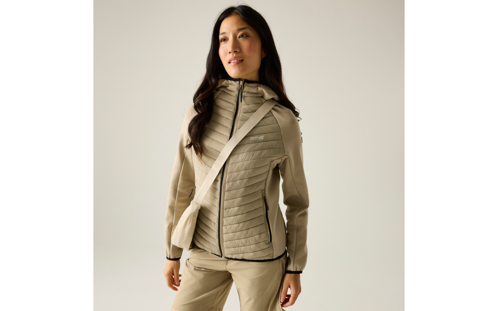 Person trägt beige gesteppte Kapuzenjacke, passende Hose und Umhängetasche; steht vor hellem einfarbigem Hintergrund, Blick leicht nach rechts