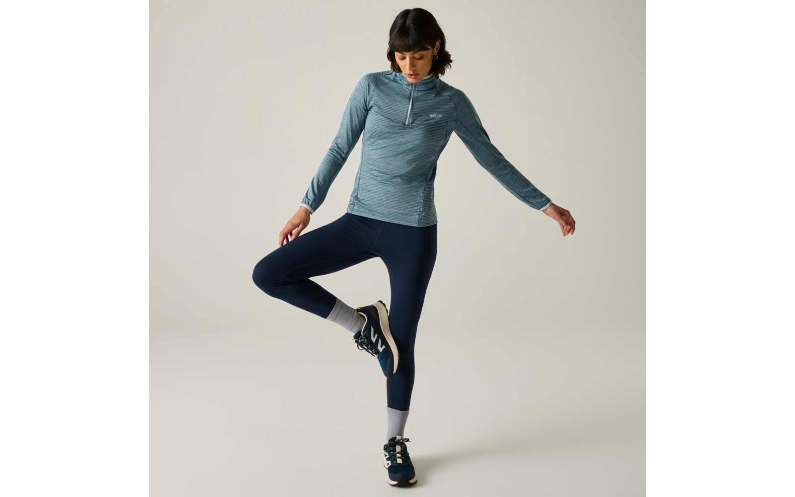 Personne aux cheveux foncés jusqu'aux épaules, haut bleu à manches longues avec courte fermeture éclair, legging bleu marine, chaussettes blanches et baskets bleues, debout sur une jambe devant un fond clair, regard vers le bas