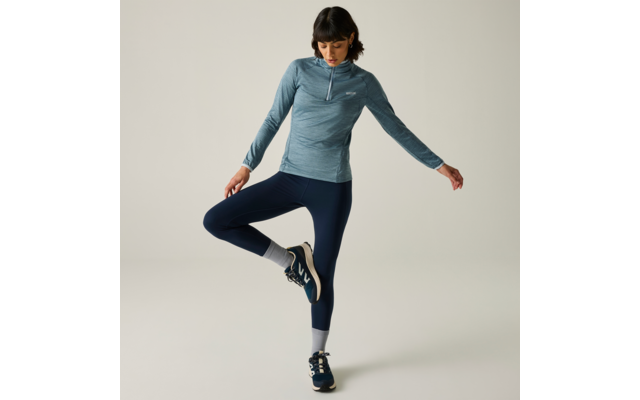 Personne aux cheveux foncés jusqu'aux épaules, haut bleu à manches longues avec courte fermeture éclair, legging bleu marine, chaussettes blanches et baskets bleues, debout sur une jambe devant un fond clair, regard vers le bas