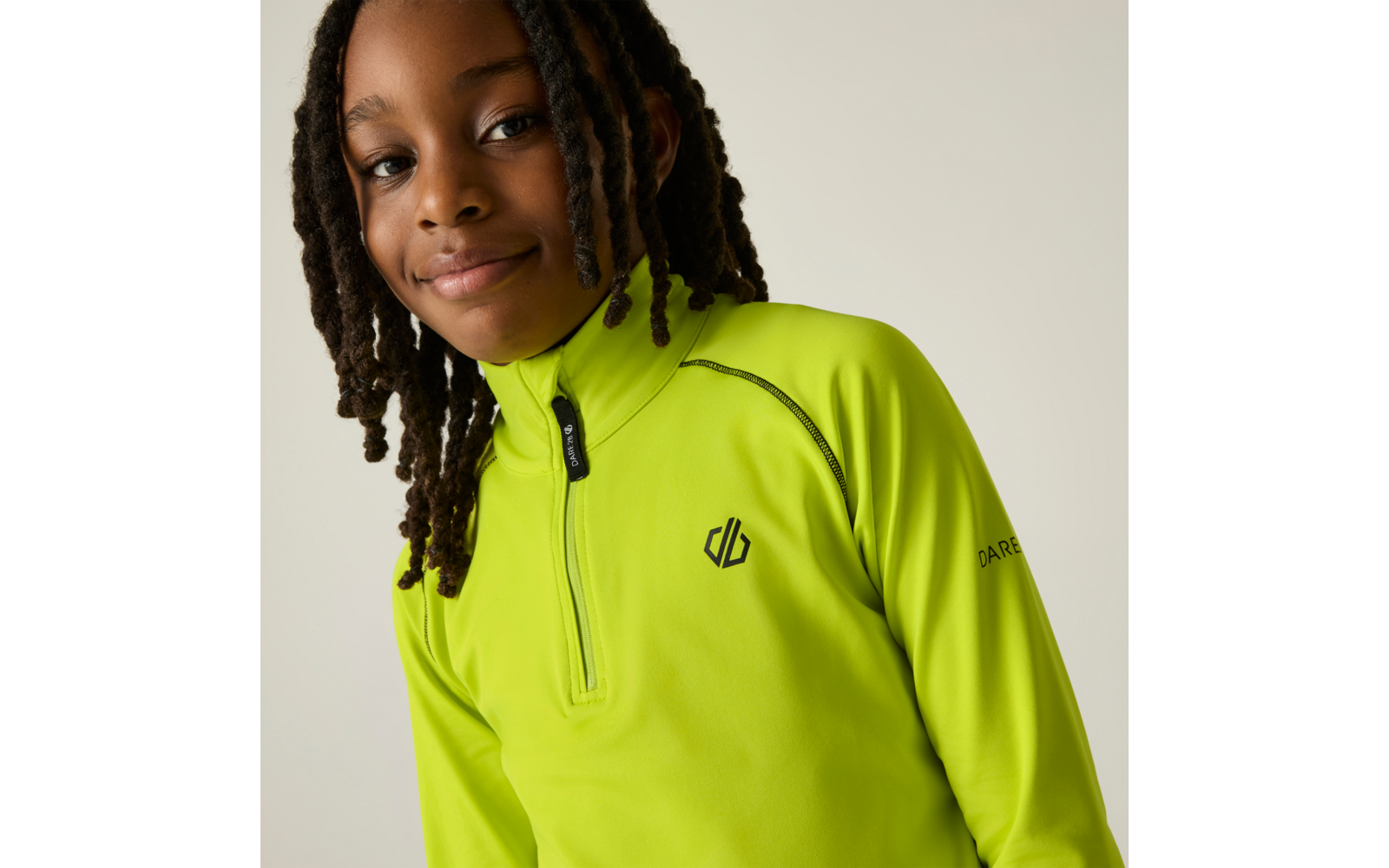Persoon met gevlochten haar draagt een neongeel-groen shirt met lange mouwen en korte rits, klein logo op de borst, tekst op de mouw, lichte effen achtergrond